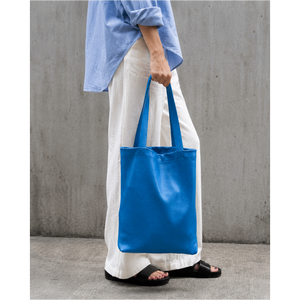 A person wearing a light blue shirt, white wide-leg pants, and black slide sandals holds a bright blue canvas tote bag against a gray concrete wall - AZURE