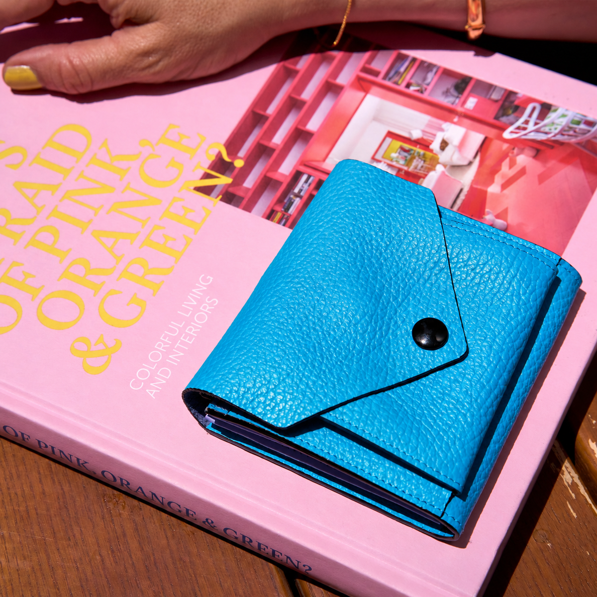 Tusk 382 Ascot Origami Wallet in Azure Italian pebble-grain cowhide leather. Closed wallet lifestyle view on a pink book with a hand visible, showing leather grain and matching stitching - AZURE