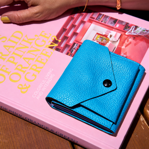 Tusk 382 Ascot Origami Wallet in Azure Italian pebble-grain cowhide leather. Closed wallet lifestyle view on a pink book with a hand visible, showing leather grain and matching stitching - AZURE
