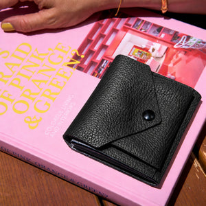 Tusk 382 Ascot Origami Wallet in black Italian pebble-grain cowhide leather. Closed wallet lifestyle view on a pink book, showing matching stitching and pebble-grain leather - BLACK