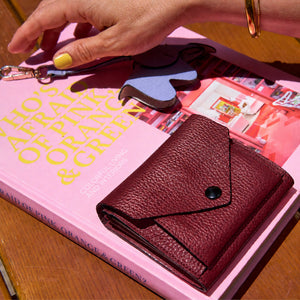 Tusk 382 Ascot Origami Wallet in oxblood Italian pebble-grain cowhide leather. Closed wallet lifestyle view on a pink book, showing matching stitching and pebble-grain leather - OXBLOOD