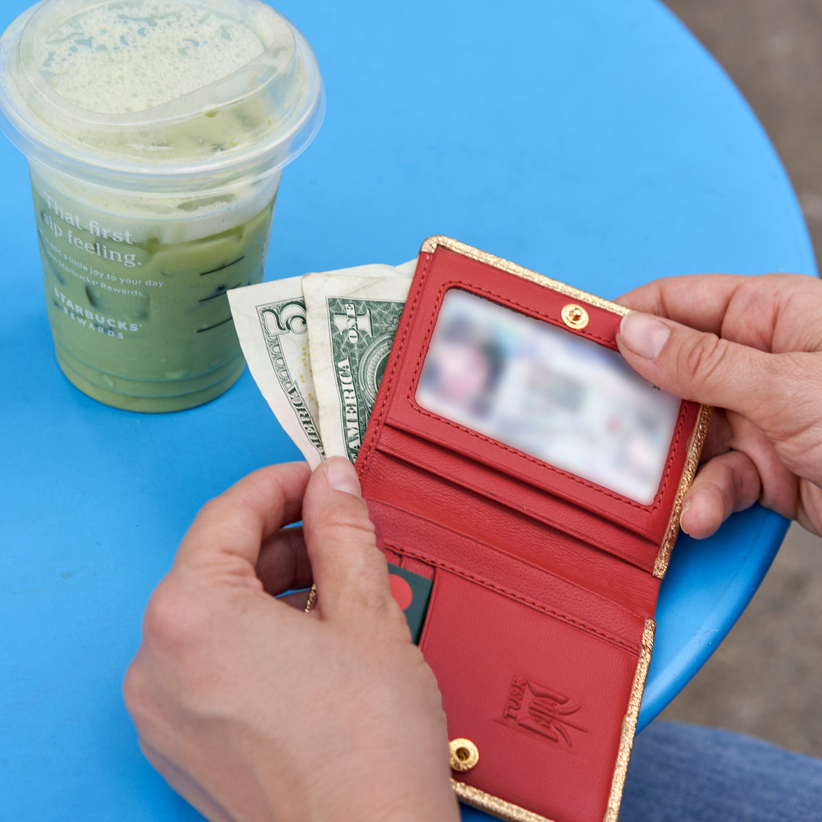 Person holding a metallic gold wallet with money and an ID card next to a green drink on a blue table - ROSE