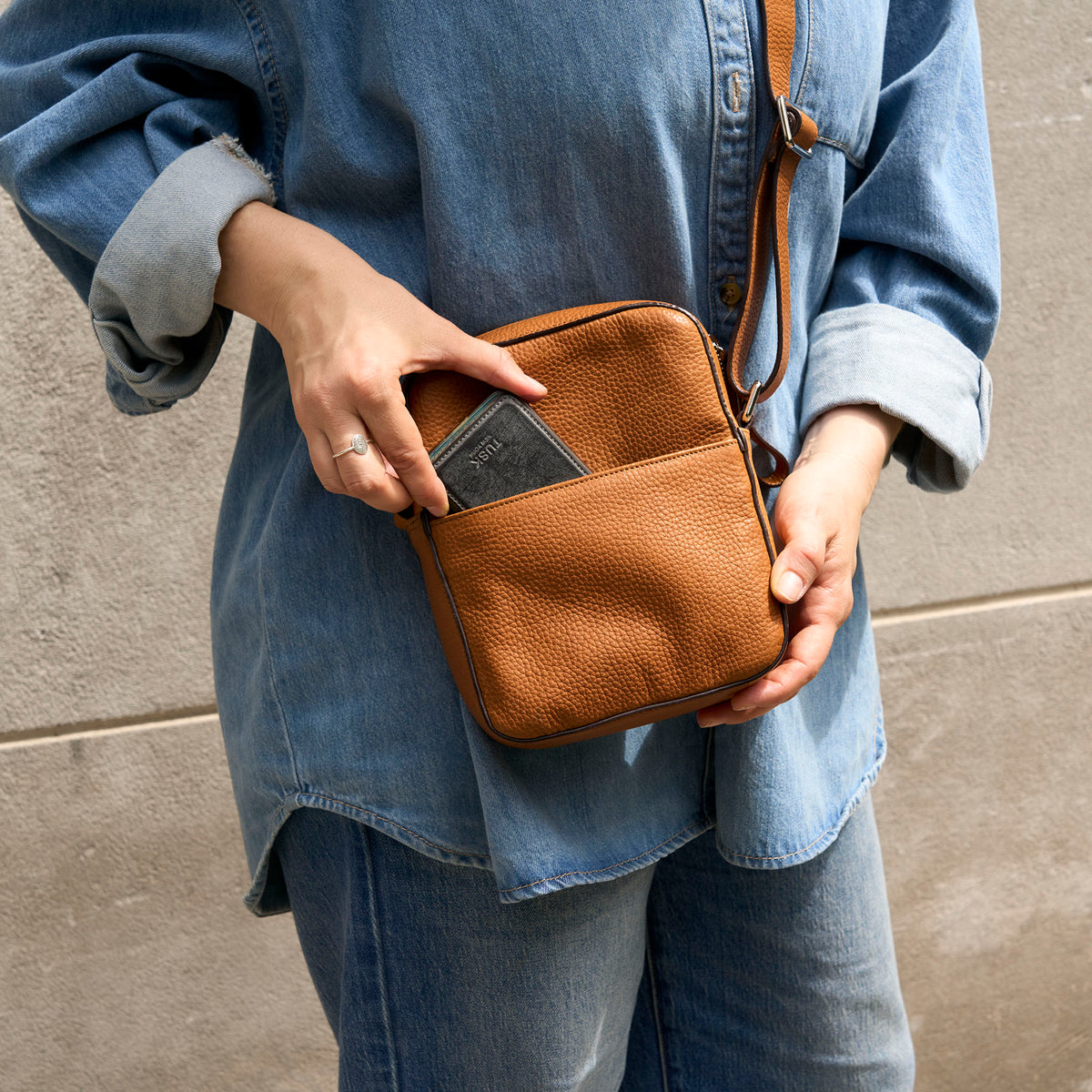 A person wearing a light blue denim shirt and jeans holds a small tan pebbled leather crossbody bag with a front slip pocket containing a gray card. The bag has a matching leather strap and is photographed against a neutral concrete wall - TOAST