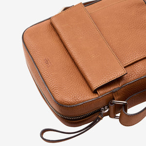 A close-up of a brown pebbled leather crossbody bag with a flap closure, zipper, and wrist strap on a light gray background - TOAST