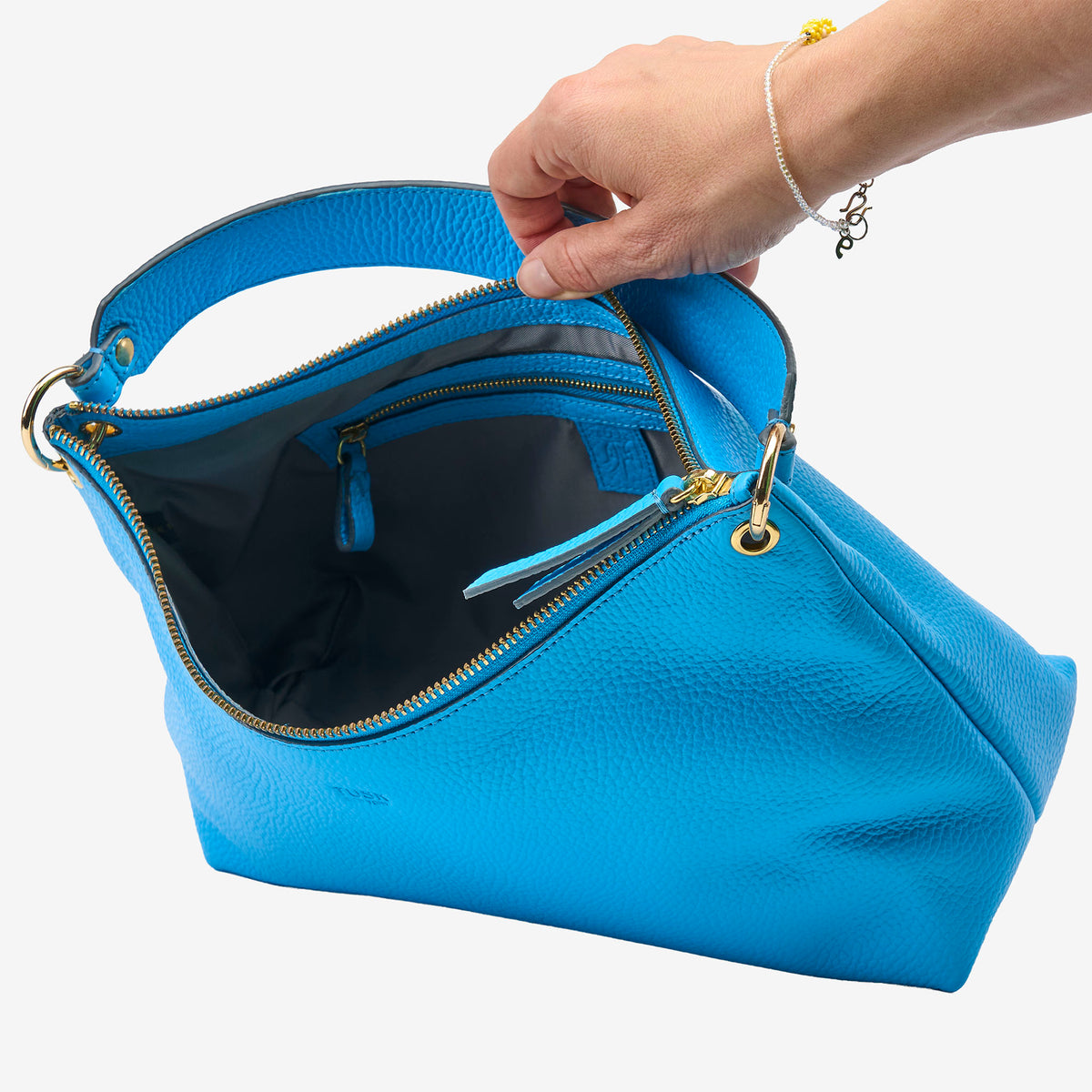 Blue handbag with gold zipper being held open by a hand on a white background - AZURE