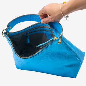 Blue handbag with gold zipper being held open by a hand on a white background - AZURE