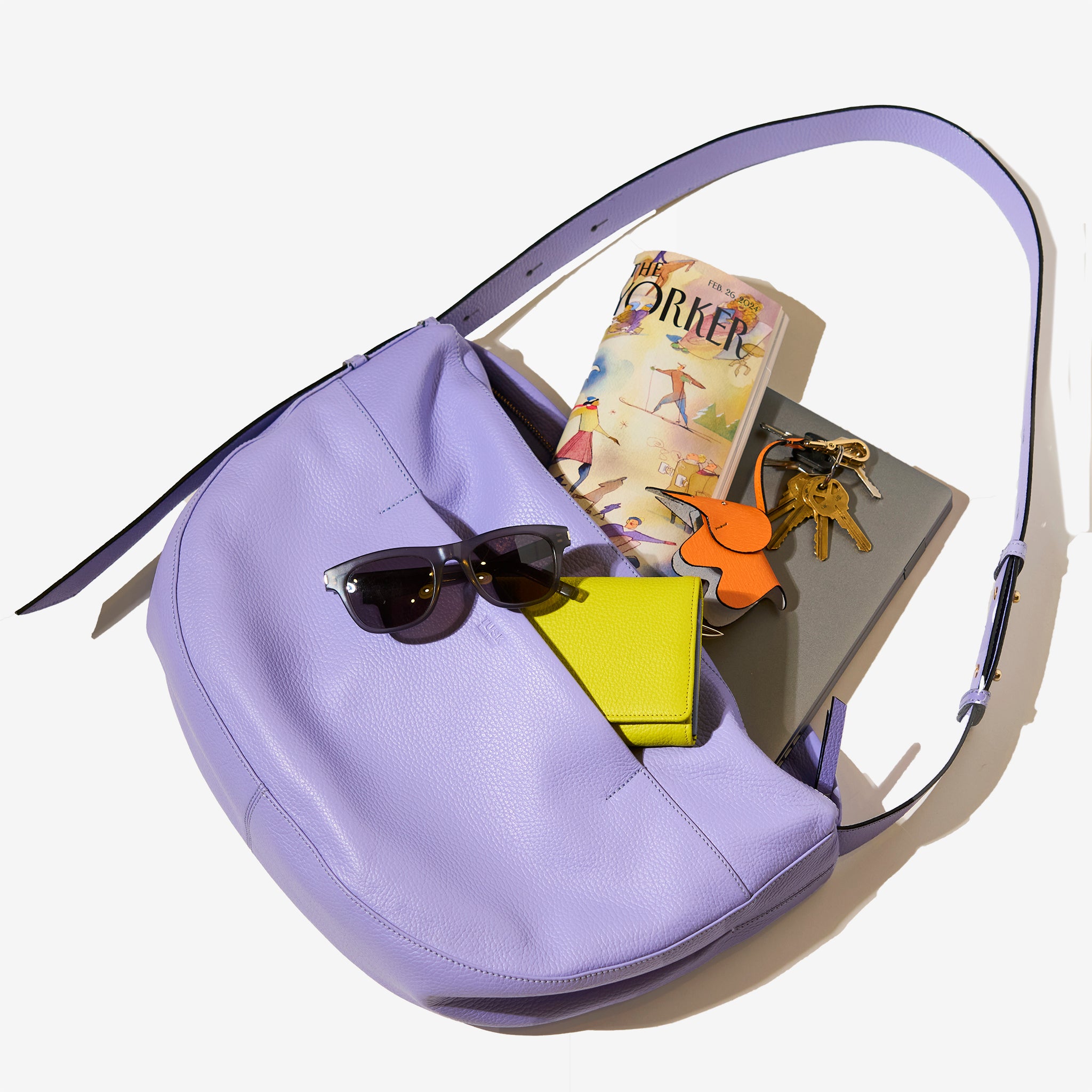 Tusk 9939 Mira Large Hobo bag in lavender-colored leather, displayed with its contents including sunglasses, a green wallet, a gray laptop, a copy of The New Yorker, and an orange leather keychain with keys, arranged on a white background - LAVENDER