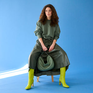 Tusk 9939 Mira Large Hobo bag in sage-colored leather, held by a woman dressed in an olive green sweatshirt and skirt, paired with neon green boots, seated on a wooden stool against a bright blue background -SAGE