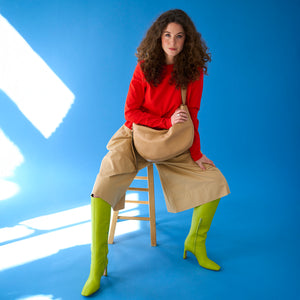 tusk-9939- woman sitting on a wooden stool wearing a red top, beige wide-leg pants, and neon green boots, holding a sand Leather Mira Large Hobo bag  against a vibrant blue background - SAND