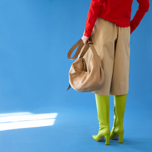 tusk 9939 back view of a woman wearing a red top, beige wide-leg pants, and neon green boots, holding a sand leatherMira Large Hobo bag by its strap against a vibrant blue background - SAND