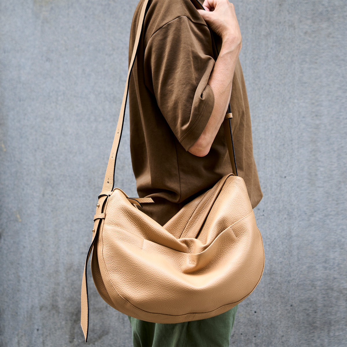 Person wearing a sand-colored leather Mira Large Hobo bag  with an adjustable strap, styled against a gray concrete wall - SAND