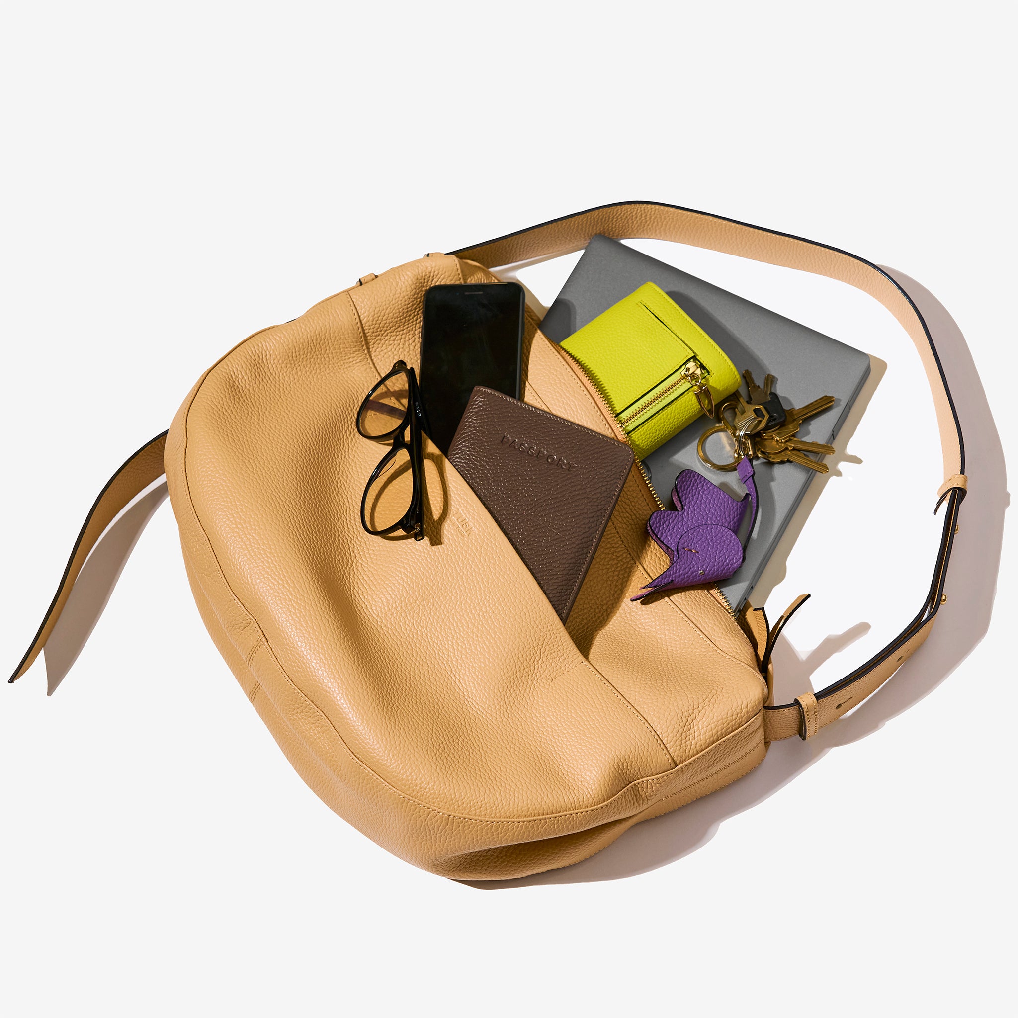 tusk 9939 sand leather Mira Large Hobo bag displayed with its contents, including glasses, a smartphone, a brown passport holder, a green wallet, a purple leather keychain, a set of keys, and a gray notebook, arranged on a white background - SAND