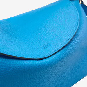 Detail of flap on blue italian leather bag with 'TUSK' branding on a light grey background - AZURE