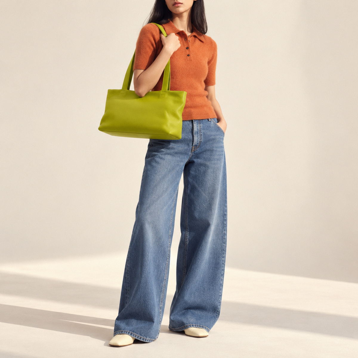 A model wearing a rust-orange short-sleeve knit polo shirt, wide-leg blue jeans, and cream pointed-toe flats, carrying a bright lime green leather tote bag against a warm off-white studio background - AVOCADO