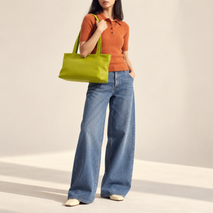 A model wearing a rust-orange short-sleeve knit polo shirt, wide-leg blue jeans, and cream pointed-toe flats, carrying a bright lime green leather tote bag against a warm off-white studio background - AVOCADO