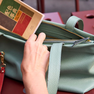 Hands placing a book into the sage green Anaya tote through its zip-top opening - SAGE